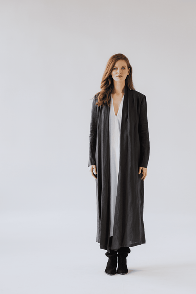 : Woman wearing a long linen jacket in natural beige tone, standing in soft natural light.
 Sieviete valkā garu lina žaketi dabīgā bēšā tonī, stāvot maigā dienasgaismā.