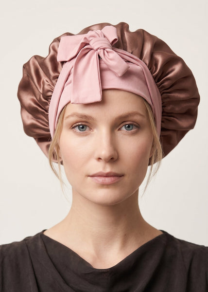 A woman wearing a silk sleep cap in two colors: a dusty pink headband with a large bow and adjustable elastic ties She is looking directly at the camera against a plain light background.