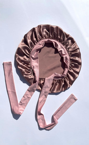 A dusty rose silk bonnet with matching ribbon ties, laid flat on a light blue background.